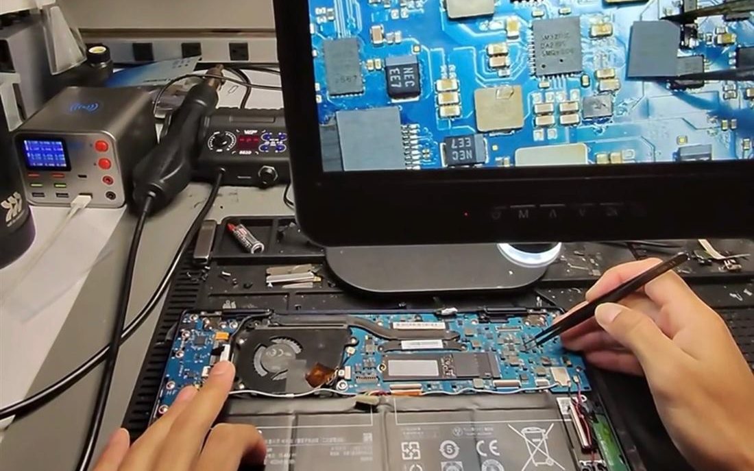 A laptop motherboard on the repair bench with an inspection microscope and rework station alongside.