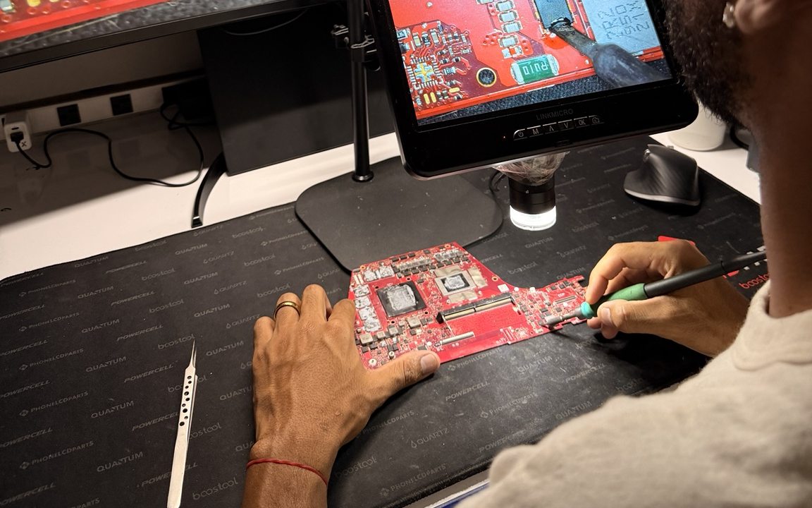 Hands working on a laptop motherboard under a digital microscope at the repair bench.